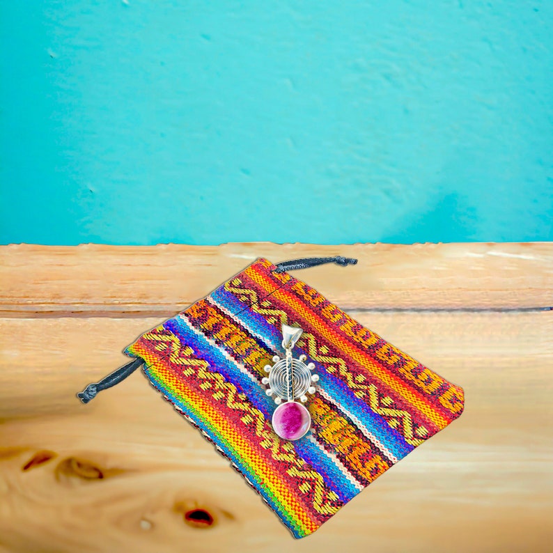 May include: A silver pendant with a pink stone and a geometric design, resting on a colorful woven pouch.