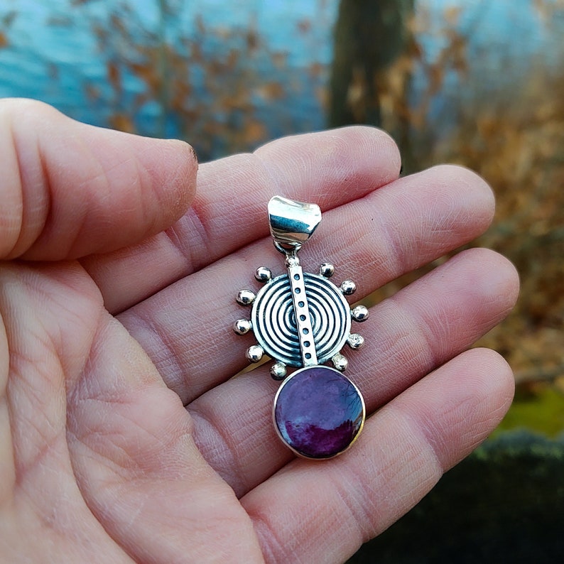 May include: A silver pendant with a circular design and a purple stone. The pendant has a silver bail for attaching to a chain.
