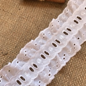 May include: White cotton eyelet lace trim with scalloped edges and small embroidered flowers. The lace is shown on a burlap background.