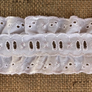 Op de afbeelding: Witte katoenen kantbies met een gegolfde rand en oogjesdetails. De kant heeft een delicaat bloemenpatroon en is perfect om een ​​toets van elegantie aan elk project toe te voegen.