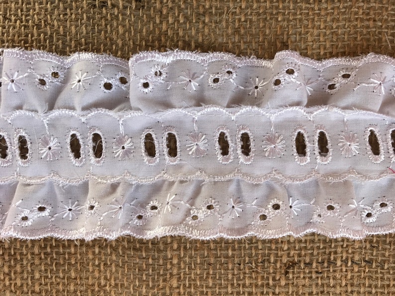 Op de afbeelding: Witte katoenen kantbies met een gegolfde rand en oogjesdetails. De kant heeft een delicaat bloemenpatroon en is perfect om een ​​toets van elegantie aan elk project toe te voegen.