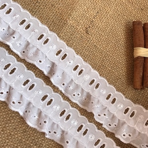 May include: White cotton eyelet lace trim with a ruffled edge. The lace has a scalloped design with small holes and embroidered flowers.