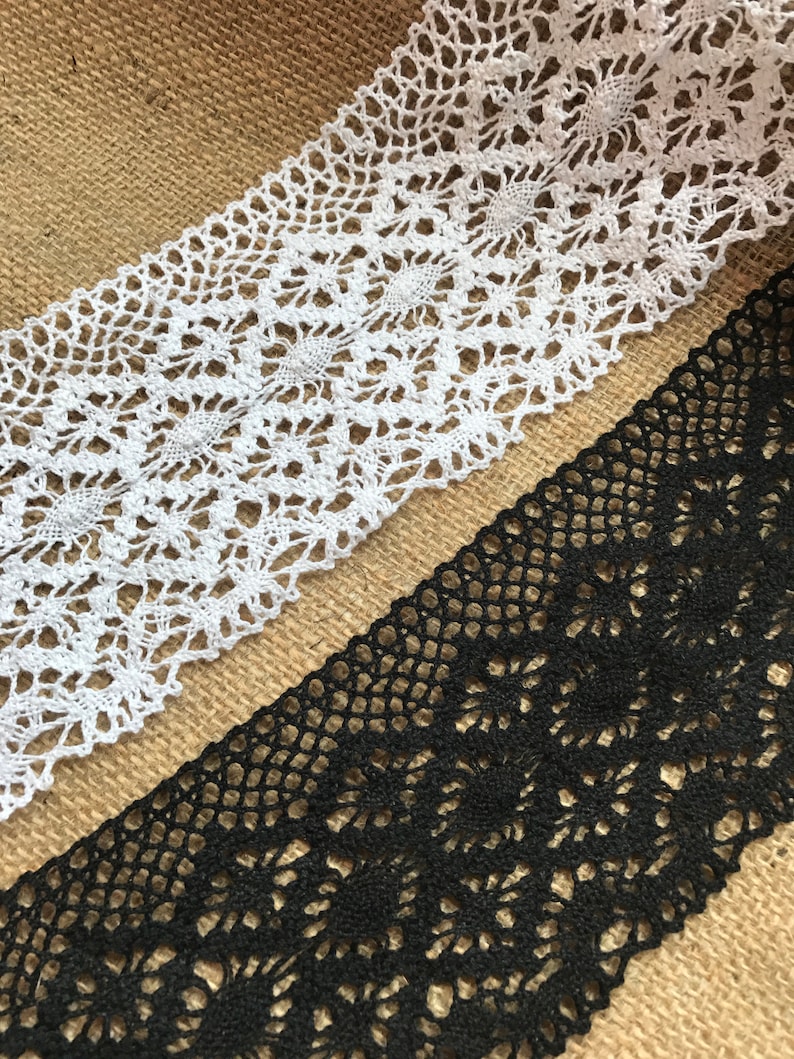 May include: Two pieces of lace trim, one white and one black, with a delicate floral pattern. The lace is laid out on a burlap background.