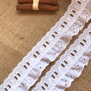 May include: White cotton lace trim with a scalloped edge and small embroidered flowers. The lace has a ruffled design and is perfect for adding a touch of elegance to any project.