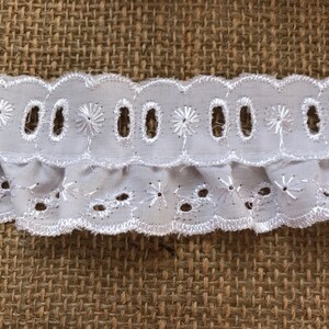 May include: White cotton lace trim with a scalloped edge and eyelet details. The lace has a ruffled bottom edge.