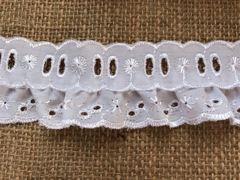 May include: White cotton lace trim with a scalloped edge and eyelet details. The lace has a ruffled bottom edge.