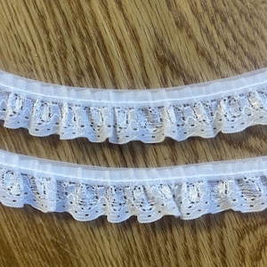 Può includere: Due strisce di passamaneria in pizzo bianco con bordo a balze. Il pizzo presenta un delicato disegno traforato con piccoli fori distanziati. La passamaneria è posta su uno sfondo di legno.