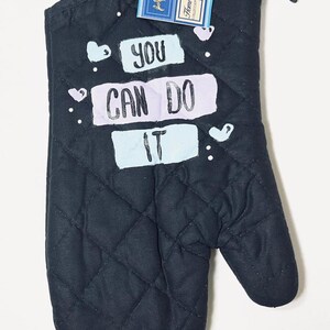 May include: A black quilted oven mitt featuring the text "YOU CAN DO IT" in white and light purple lettering, accented with small white hearts. A product tag attached to the top indicates dimensions of 7 inches by 13 inches.