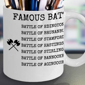 May include: White ceramic mug with a black handle. The mug has a black graphic that says "Famous Bat'" and lists a series of battles: Battle of Edington, Battle of Brunanbu, Battle of Stamfori, Battle of Hastings, Battle of Stirling, Battle of Bannock, Battle of Agincour.