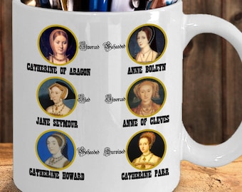May include: White ceramic mug with a design of six women, each in a gold oval frame. The women are identified as the six wives of King Henry VIII: Catherine of Aragon, Anne Boleyn, Jane Seymour, Anne of Cleves, Catherine Howard, and Catherine Parr. Each woman's name is printed below her portrait. The words "Divorced", "Beheaded", "Died", "Divorced", "Beheaded", and "Survived" are printed above the portraits.
