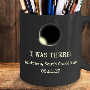 May include: A black ceramic mug filled with pens and markers. The mug has a handle and features the text "I WAS THERE" with the location "Andrews, South Carolina" and the date "08.21.17". A graphic of a solar eclipse is also on the mug.
