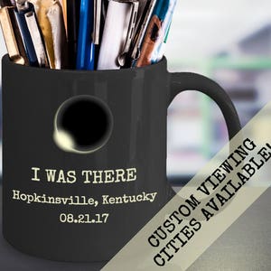 May include: Black ceramic mug with a white silhouette of a solar eclipse and the text "I Was There Hopkinsville, Kentucky 08.21.17". The mug has a white handle and is filled with pens and pencils.