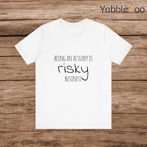 May include: White t-shirt with the text "BEING AN ACTUARY IS risky BUSINESS" in black. The shirt is made of a soft material and has short sleeves. The text is centered on the front of the shirt.