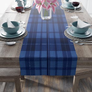 May include: Blue and black plaid table runner with a woven texture. The runner is laid out on a wooden table with place settings.