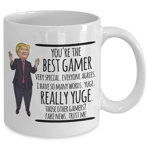 May include: White ceramic coffee mug with a cartoon image of a man in a suit and red tie. The text on the mug reads "You're the Best Gamer Very Special. Everyone Agrees. I have so many words. YUGE. REALLY YUGE. Those other gamers? Fake news. Trust me."