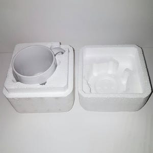 K&ouml;nnte beinhalten: Eine wei&szlig;e Keramiktasse mit Henkel, verpackt in einem wei&szlig;en Styropor-Karton mit einem separaten Styropor-Einsatz f&uuml;r die Tasse.