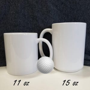 May include: Two white ceramic mugs with handles. The mug on the left is 11 ounces and the mug on the right is 15 ounces. A white golf ball is in front of the mugs.