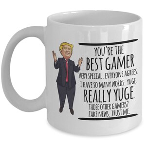 May include: White ceramic coffee mug with a cartoon image of a man in a suit and red tie. The text on the mug reads "You're the Best Gamer. Very special. Everyone agrees. I have so many words. Yuge. Really Yuge. Those other gamers? Fake news. Trust me."