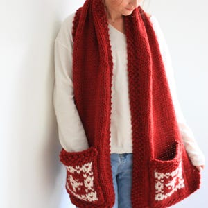 May include: A red knitted scarf with two white knit pockets. The scarf is worn by a person with long brown hair.