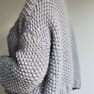 May include: A gray knitted sweater with a loose fit and long sleeves. The sweater is made of a chunky yarn and has a ribbed texture.