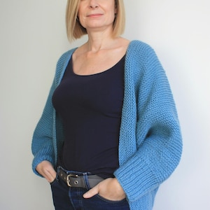 May include: A woman wearing a light blue knitted cardigan over a black tank top and blue jeans. She is standing with her hands in her pockets.