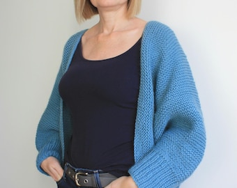 May include: A woman wearing a light blue knitted cardigan over a black vest top and blue jeans. She is standing with her hands in her pockets.