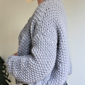 May include: A light gray knitted sweater with a relaxed fit and long sleeves. The sweater has a slightly cropped length and a ribbed hem.