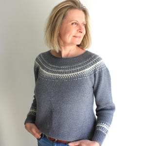 May include: A grey knitted jumper with a white and grey patterned design around the neckline and cuffs. The jumper is worn by a person with blonde hair and is paired with blue jeans.