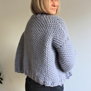 May include: A light gray knitted cardigan with a slightly cropped length. The cardigan is made with a textured stitch and has long sleeves.