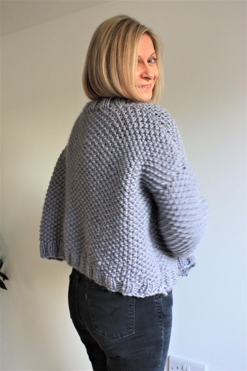 May include: A light gray knitted cardigan with a slightly cropped length. The cardigan is made with a textured stitch and has long sleeves.