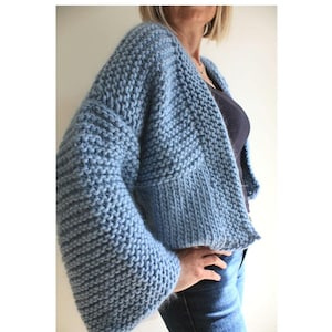 Może przedstawiać: Jas w kolorze jasnoniebieskim, wykonany z dzianiny, o luźnym kroju i długich rękawach.  Jas ma dekolt w kształcie litery V i krótki krój.