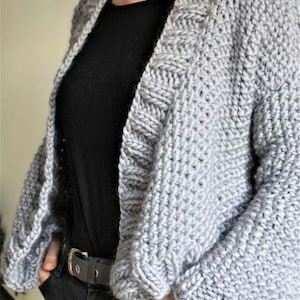 May include: A gray knitted cardigan with a loose fit and a V-neck. The cardigan has a textured stitch pattern and is shown being worn over a black shirt.