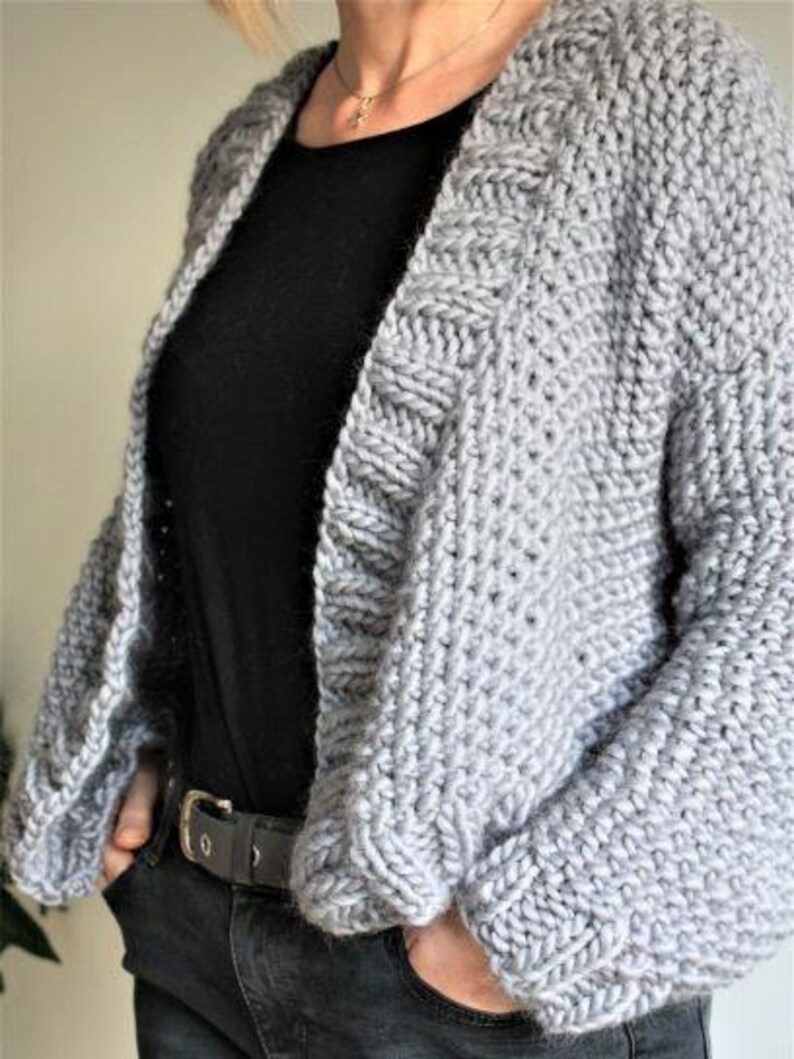 May include: A gray knitted cardigan with a loose fit and a V-neck. The cardigan has a textured stitch pattern and is shown being worn over a black shirt.
