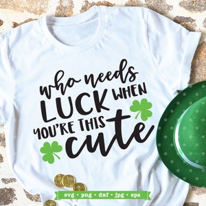 May include: White t-shirt with black text that reads "who needs luck when you're this cute" with a green shamrock on either side of the text.  The shirt is laying on a brown brick surface with gold coins and a green shamrock hat.