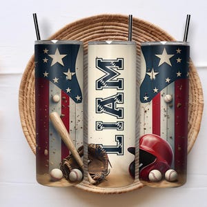May include: Three baseball-themed tumblers with black straws. The tumblers feature an American flag design with stars and stripes, baseballs, a bat, a glove, and a red helmet. The center tumbler has the name "LIAM" in navy blue.