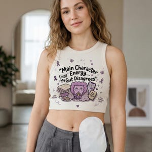 Könnte beinhalten: Cremefarbenes Crop-Top mit dem Text "Main Character Until Energy... My Gut Disagrees." Das Top zeigt eine Cartoon-Löwen-Illustration und andere Grafiken. Das Top ist ärmellos und hat einen runden Ausschnitt.