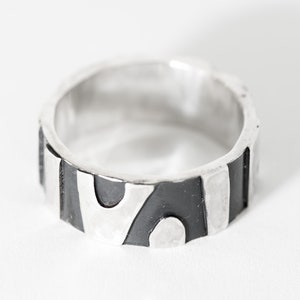 May include: A silver ring with a black and white abstract design. The ring is made of metal and has a smooth finish.