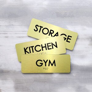 K&ouml;nnte beinhalten: Drei goldene Schilder aus Metall mit schwarzem Text. Die Schilder zeigen "STORAGE", "KITCHEN" und "GYM".