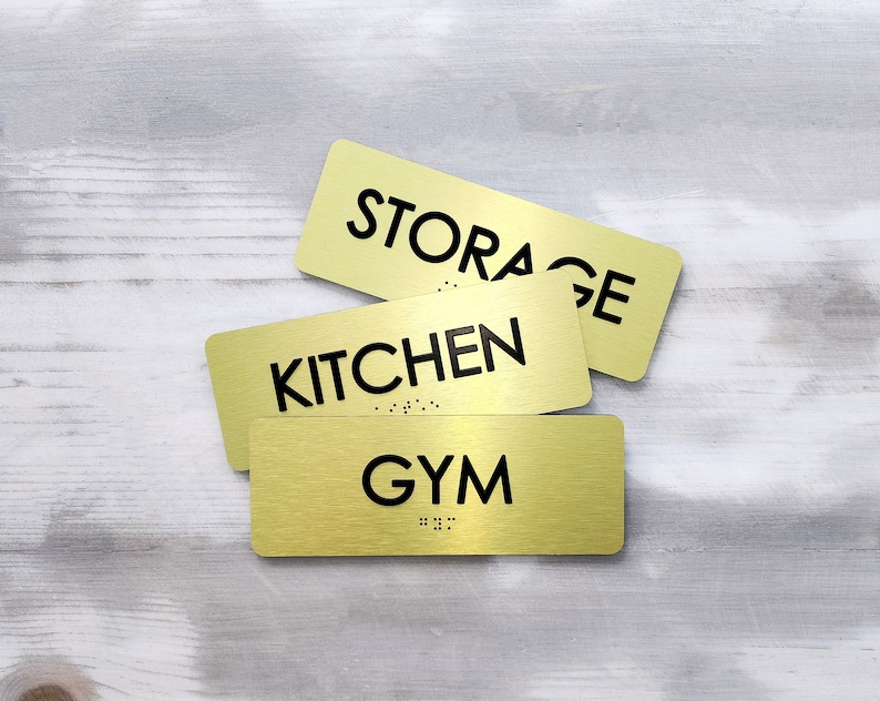 K&ouml;nnte beinhalten: Drei goldene Schilder aus Metall mit schwarzem Text. Die Schilder zeigen "STORAGE", "KITCHEN" und "GYM".