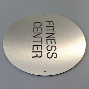 May include: A circular, brushed metal sign displaying the words "FITNESS CENTER" cut out vertically. The sign has a black border and two small holes. The background is a light grey.