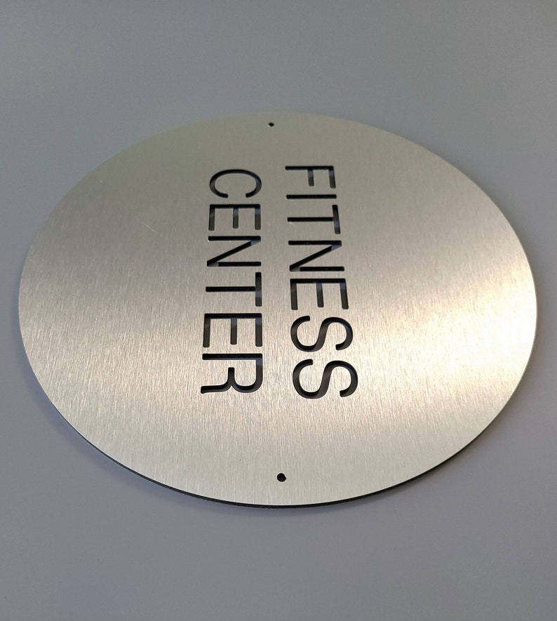 May include: A circular, brushed metal sign displaying the words "FITNESS CENTER" cut out vertically. The sign has a black border and two small holes. The background is a light grey.