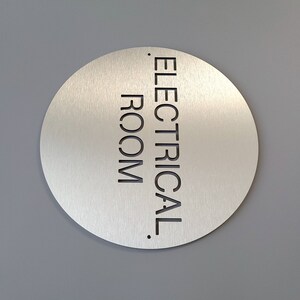 May include: A circular, brushed metal sign with the words "ELECTRICAL ROOM" in black, sans-serif font. The sign is mounted on a grey wall.