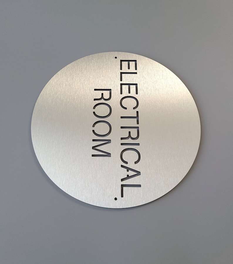 May include: A circular, brushed metal sign with the words "ELECTRICAL ROOM" in black, sans-serif font. The sign is mounted on a grey wall.