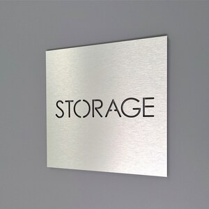 May include: A square, brushed metal sign with the word "STORAGE" cut out in a sans-serif font. The sign is mounted on a gray wall. The sign is angled slightly, showing its thickness and the texture of the metal.