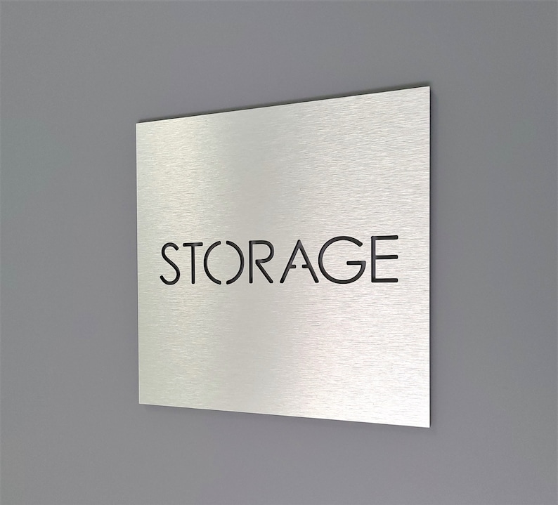 May include: A square, brushed metal sign with the word "STORAGE" cut out in a sans-serif font. The sign is mounted on a gray wall. The sign is angled slightly, showing its thickness and the texture of the metal.