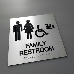 Pu&ograve; includere: Un cartello argentato con testo nero e icone che recita "FAMILY RESTROOM" con braille sotto. Le icone includono un uomo, una donna, una sedia a rotelle e un bambino.