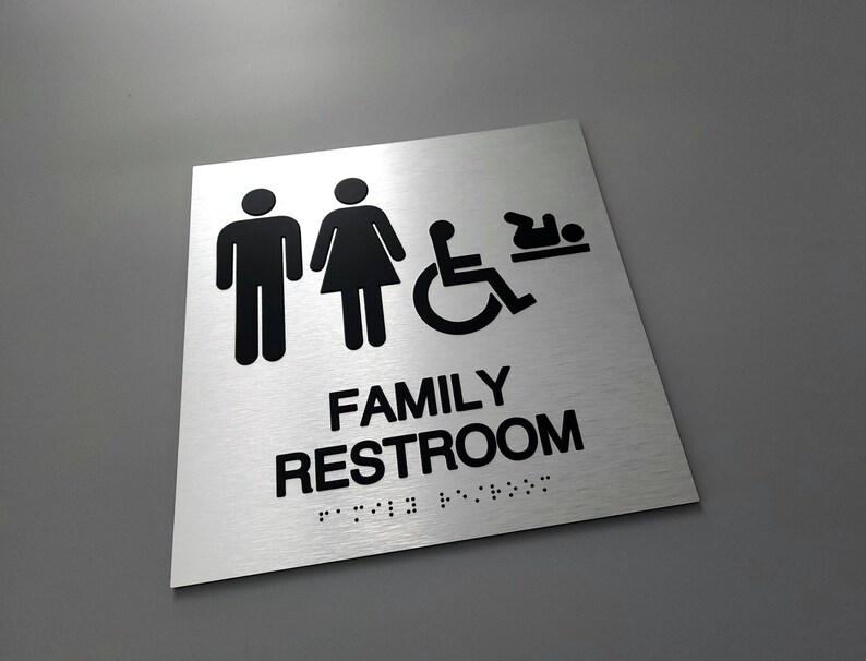 Pu&ograve; includere: Un cartello argentato con testo nero e icone che recita "FAMILY RESTROOM" con braille sotto. Le icone includono un uomo, una donna, una sedia a rotelle e un bambino.