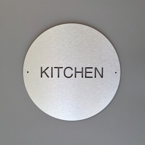 May include: A circular, brushed metal sign with the word "KITCHEN" cut out in black, centrally positioned. Two small dots flank the text. The sign is mounted on a grey wall.