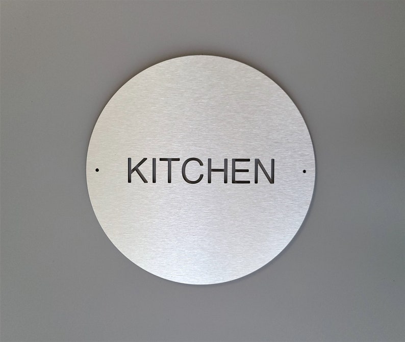 May include: A circular, brushed metal sign with the word "KITCHEN" cut out in black, centrally positioned. Two small dots flank the text. The sign is mounted on a grey wall.