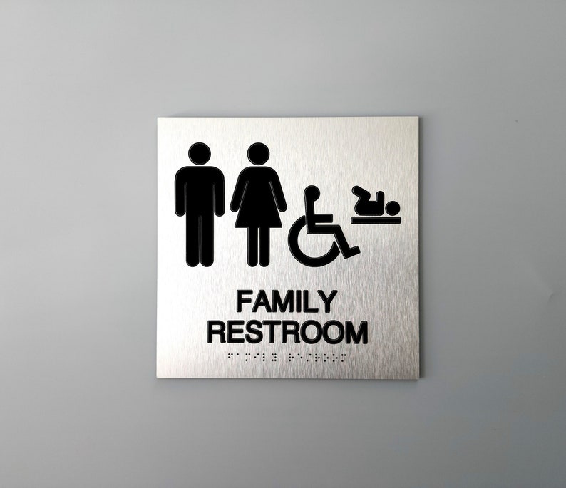 Pu&ograve; includere: Un cartello in metallo argentato con testo nero e icone. Il cartello recita "FAMILY RESTROOM" con icone di un uomo, una donna, una persona su sedia a rotelle e un bambino. Il cartello ha anche testo in Braille.